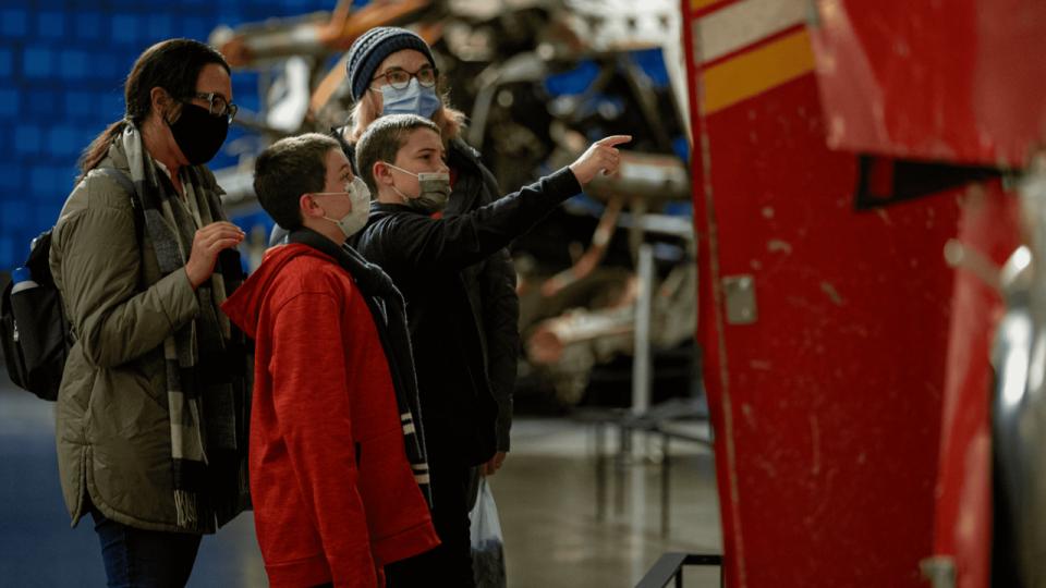 Two young boys and two women observe FDNY Ladder 3 in Memorial Hall of the 9/11 Memorial Museum.  One of the boys gestures toward the front of the red fire truck as the damaged rear can be observed in the background.