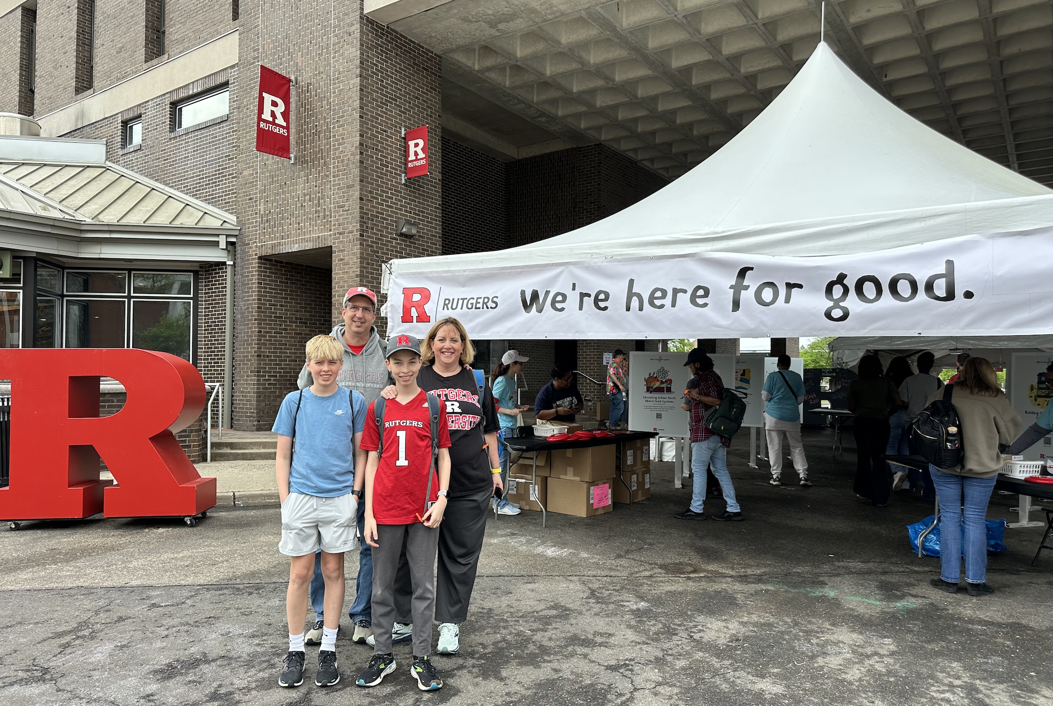 A family in Rutgers Day apparel standing in front of the "Here for Good" tent
